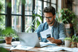 © btiger - Focused Male Entrepreneur in Glasses Using Mobile and Laptop with Documents at Desk by Window
