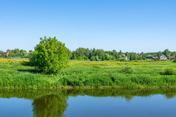 Wall Mural - Summer countryside landscape with village and river under blue sky. Rural river with green meadow peaceful landscape.