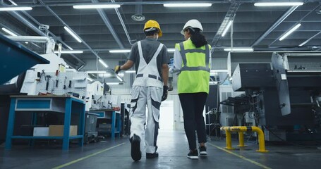 Poster - Two Multiethnic Employees Walking in an Industrial Factory Hall. Female Engineer Using a Tablet, Explaining the Scope of Work to a Young Technician. Workers Wearing Hard Hats for Health and Safety