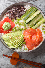 Canvas Print - Asian sushi bowl with salmon, rice, avocado, cucumber, green onion, seaweed, sesame, pickled ginger and wasabi close-up on the table. Vertical top view from above