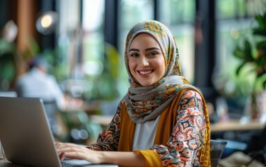 Wall Mural - A smiling Muslim woman in a hijab works on her laptop in a cafe, surrounded by the warm, inviting atmosphere of a bustling coffee shop