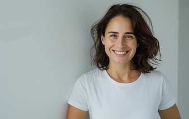 Wall Mural - A smiling brunette woman with short, wavy hair wears a white t-shirt and stands in front of a light grey wall