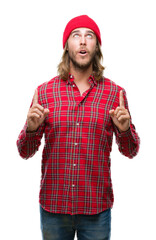 Canvas Print - Young handsome man with long hair wearing red cap over isolated background amazed and surprised looking up and pointing with fingers and raised arms.