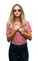 Poster - Young beautiful blonde woman wearing sunglasses over isolated background smiling with hands on chest with closed eyes and grateful gesture on face. Health concept.