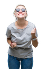 Poster - Young blonde woman wearing sunglasses over isolated background very happy and excited doing winner gesture with arms raised, smiling and screaming for success. Celebration concept.