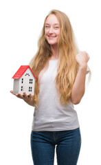 Sticker - Blonde teenager woman holding family house screaming proud and celebrating victory and success very excited, cheering emotion