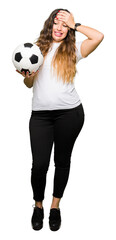 Poster - Young adult woman holding soccer football ball stressed with hand on head, shocked with shame and surprise face, angry and frustrated. Fear and upset for mistake.