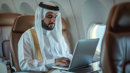 emirati man in traditional thobe and ghutra sitting on airplane seat working with laptop computer focused typing light interior plane