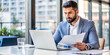 © hobonski - Focused Businessman Reviewing Documents at Office Desk. A serious businessman in a suit intently reviews documents while working on his laptop in a bright, modern office.
