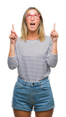 Poster - Young beautiful woman wearing glasses over isolated background amazed and surprised looking up and pointing with fingers and raised arms.