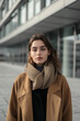 © byerenyerli - A woman wearing a stylish camel coat and a cozy scarf stands in front of a modern building.