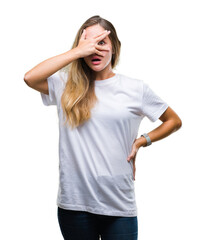 Canvas Print - Young beautiful blonde woman wearing casual white t-shirt over isolated background peeking in shock covering face and eyes with hand, looking through fingers with embarrassed expression.