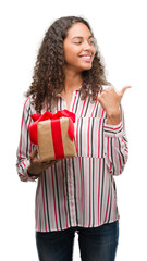 Canvas Print - Young hispanic woman in love holding present pointing and showing with thumb up to the side with happy face smiling