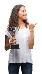 Canvas Print - Young hispanic woman holding trophy pointing and showing with thumb up to the side with happy face smiling