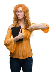 Poster - Young redhead student woman holding a book with surprise face pointing finger to himself