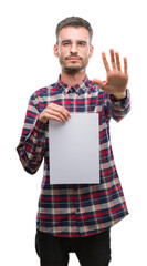 Poster - Young hipster adult man holding blank paper sheet with open hand doing stop sign with serious and confident expression, defense gesture