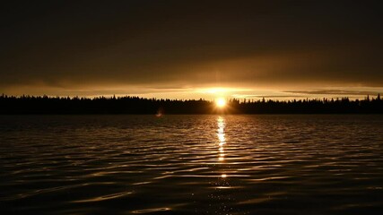 Wall Mural - Time lapse of an early morning clip of a lake with small waves reflecting the morning gold colored glow as the sun rises through distant spruce trees. The sky is filled with ominous looking clouds.

