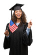 © Krakenimages.com - Young hispanic woman wearing graduation uniform holding flag of United States very happy pointing with hand and finger to the side
