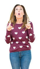 Wall Mural - Beautiful middle age woman wearing hearts sweater over isolated background amazed and surprised looking up and pointing with fingers and raised arms.