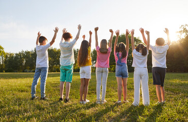 Wall Mural - Group of kids friends raising hands up on green grass in the park standing back in a line at sunset. Children having fun together outdoors on a sunny summer day in casual clothes in nature.