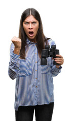 Wall Mural - Young hispanic woman holding binoculars annoyed and frustrated shouting with anger, crazy and yelling with raised hand, anger concept