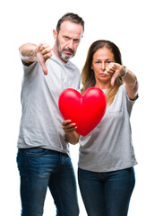 Canvas Print - Middle age hispanic casual couple in love holding red heart over isolated background with angry face, negative sign showing dislike with thumbs down, rejection concept