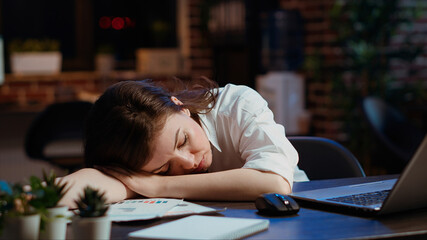 Sleepy employee asleep on computer desk late at night in brick wall office. Panning shot of tired businesswoman sleeping with head on table while working overnight on laptop, camera B