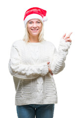 Wall Mural - Young beautiful blonde woman wearing christmas hat over isolated background with a big smile on face, pointing with hand and finger to the side looking at the camera.