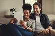 © peopleimages.com - Interracial couple, tablet and smile in home for media, streaming and social network on sofa. Happy man, woman or technology on couch for TV show, entertainment or ecommerce in living room on website