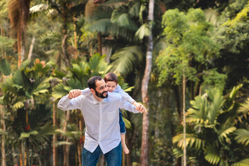 A joyful Brazilian father carrying his young son on his back in a lush, green forest. They are laughing and playing, highlighting a special moment of father-son bonding on Father's Day	