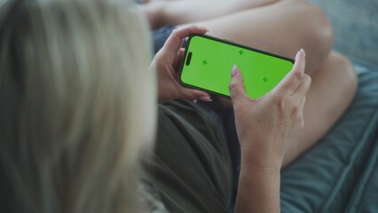 Poster - back view of woman holding mobile phone with green screen and watching video content lying on sofa i