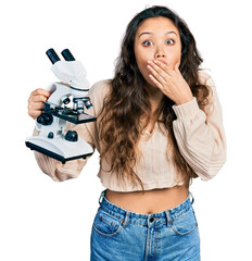 Sticker - Young hispanic girl holding microscope covering mouth with hand, shocked and afraid for mistake. surprised expression