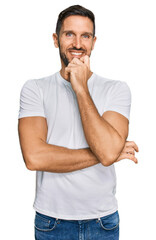 Handsome man with beard wearing casual white t shirt looking confident at the camera with smile with crossed arms and hand raised on chin. thinking positive.