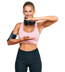 Wall Mural - Young blonde woman wearing gym clothes and using headphones gesturing with hands showing big and large size sign, measure symbol. smiling looking at the camera. measuring concept.