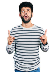 Sticker - Young arab man with beard wearing casual striped sweater amazed and surprised looking up and pointing with fingers and raised arms.