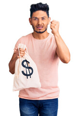 Wall Mural - Handsome latin american young man holding money bag with dollar symbol annoyed and frustrated shouting with anger, yelling crazy with anger and hand raised