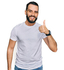 Young man with beard wearing casual white t shirt doing happy thumbs up gesture with hand. approving expression looking at the camera showing success.