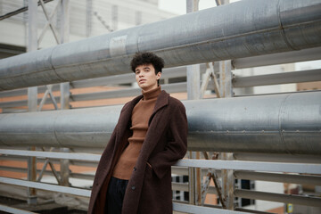 Wall Mural - Fashionable young man in a brown coat and turtleneck sweater leaning against a metal railing in front of a large structure