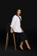 Poster - Beautiful young woman with stylish accessories sitting on chair against black background