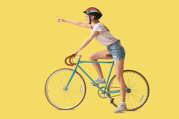 Poster - Beautiful young woman in helmet with bicycle pointing at something on yellow background