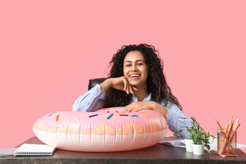 Sticker - Happy female office worker with inflatable ring sitting at workplace against pink background. Summer vacation concept