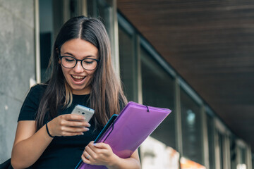 Wall Mural - Girl university student user holding cell phone using mobile applications tech on smartphone looking at cellphone, chatting, texting, checking social media, educational apps standing outside campus.