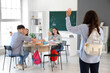 © Pixel-Shot - Female teenage student with backpack waving hand to her classmates in classroom, back view