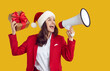 © Studio Romantic - Happy saleswoman advertising holiday sale. Woman in red suit and Santa hat standing isolated on yellow studio background, holding giftbox and yelling through megaphone. Christmas and New Year concept