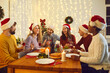© Studio Romantic - Friends communicate with each other sitting at festive table and enjoying Christmas Eve. Young multiracial people in Santa hats tell funny stories sitting at home in room with Christmas decorations.