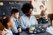© Seventyfour - Happy female teacher of Black ethnicity using laptop explaining new theme to diverse group of kids around her in elective class