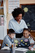 © Seventyfour - Vertical shot of smiling female teacher of Black ethnicity talking to primary students looking at laptop screen