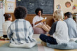 © Seventyfour - Young female teacher of Black ethnicity in bean bag chair reading book together with children sharing thoughts about story while sitting on floor in classroom