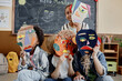 © Seventyfour - Group of four kids in classroom holding handmade cardboard abstract faces presenting art project results