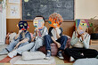 © Seventyfour - Group of unrecognizable primary schoolers sitting together in bean bag chair and on classroom floor hiding faces behind cardboard mask crafts in abstract style